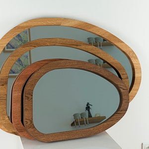 Op de afbeelding: Een set van drie gelaagde, organisch gevormde spiegels met opvallende houten frames. De frames vertonen variërende tinten van natuurlijke houtnerven, variërend van licht eiken tot een diepere, rijkere bruine tint in een gestapelde opstelling.