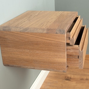 Può includere: Un comodino sospeso a parete realizzato in legno naturale con venature a vista. L'unità è dotata di due cassetti sovrapposti con maniglie orizzontali integrate lungo i bordi anteriori e angoli arrotondati sulla superficie superiore.