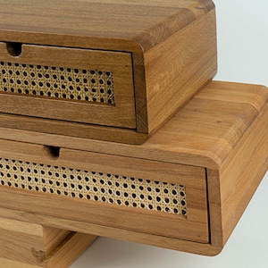 May include: Two stacked wooden storage boxes with rounded edges and a natural brown finish. Each box features a single drawer with a front panel made of woven light beige cane webbing and a small semi-circular cutout for opening the drawer.