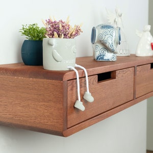 Op de afbeelding: Een zwevend houten plank met twee laden, gebeitst in een warme bruine tint. De plank is versierd met decoratieve items, waaronder een kleine potplant, een grillige keramische pot met bungelende benen en keramische beeldjes.