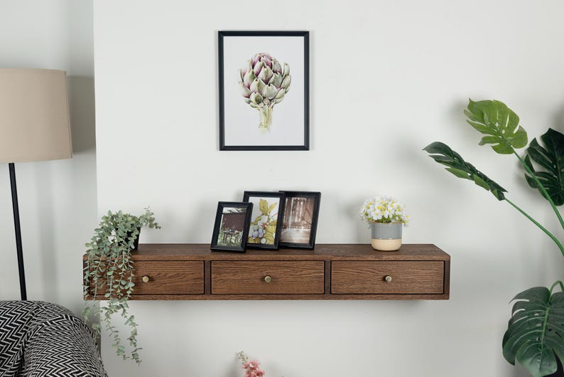 Puede incluir: Una estanter&iacute;a flotante de color marr&oacute;n oscuro con tres cajones y pomos de lat&oacute;n. Encima de la estanter&iacute;a hay tres cuadros enmarcados y una impresi&oacute;n bot&aacute;nica enmarcada de una alcachofa. Se ven una peque&ntilde;a planta en maceta y una l&aacute;mpara.