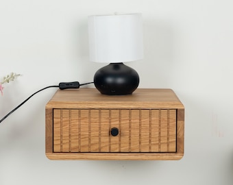 May include: A wall-mounted light oak bedside table featuring a single drawer with a vertical slatted front and a small black circular knob. A black table lamp with a white cylindrical shade sits on top of the wooden floating nightstand.