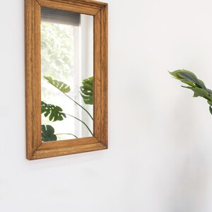 May include: A rectangular wooden framed mirror with a light brown finish. The mirror reflects a window with green foliage outside.