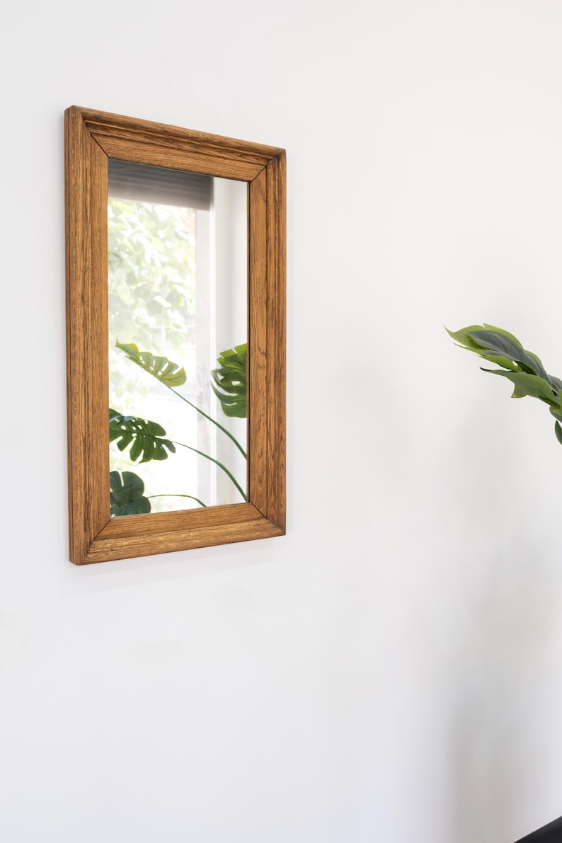 May include: A rectangular wooden framed mirror with a light brown finish. The mirror reflects a window with green foliage outside.
