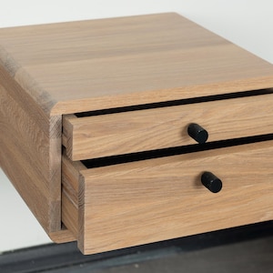 May include: A light wood floating nightstand with two drawers and black knobs. The nightstand is mounted to a white wall.
