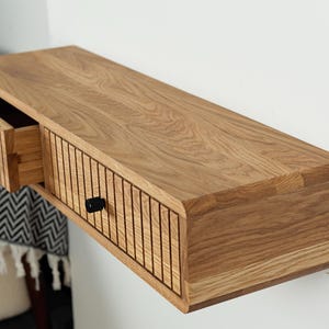 Puede incluir: Una estantería de pared de madera con dos cajones. Los cajones tienen un frente de listones y pomos negros. La estantería es de madera marrón claro.