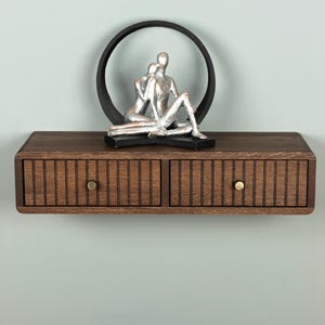 Op de afbeelding: Een zwevende wandplank van donkerbruin hout met twee lades voorzien van verticale latten en kleine ronde koperen knoppen. Bovenop de plank staat een decoratief beeld van twee figuren die elkaar omhelzen in een zwart cirkelvormig frame.