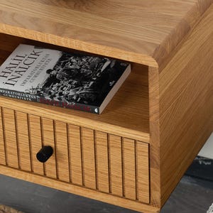 May include: A wall-mounted light oak nightstand featuring a single drawer with vertical slats and a small black circular knob. An open shelf above the drawer holds a book titled Halil Inalcik Imparatorluktan Cumhuriyete with a black and white cover.