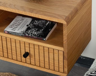 May include: A wall-mounted light oak nightstand featuring a single drawer with vertical slats and a small black circular knob. An open shelf above the drawer holds a book titled Halil Inalcik Imparatorluktan Cumhuriyete with a black and white cover.