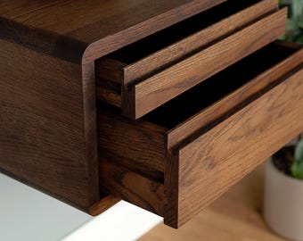 Può includere: Una mensola sospesa a parete realizzata in legno marrone scuro con una venatura prominente. Il mobile presenta due cassetti sovrapposti con maniglie a incasso orizzontali integrate lungo i bordi superiori e angoli laterali arrotondati.