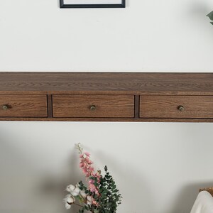 Puede incluir: Una estanter&iacute;a flotante de madera con tres cajones. La estanter&iacute;a es de color marr&oacute;n c&aacute;lido y tiene pomos de color lat&oacute;n. Los cajones est&aacute;n espaciados uniformemente. La estanter&iacute;a est&aacute; montada en una pared blanca.