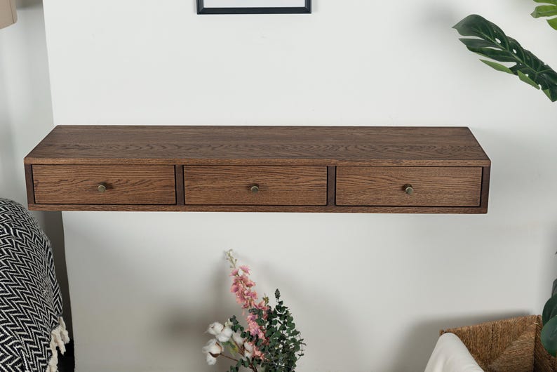 Puede incluir: Una estanter&iacute;a flotante de madera con tres cajones. La estanter&iacute;a es de color marr&oacute;n c&aacute;lido y tiene pomos de color lat&oacute;n. Los cajones est&aacute;n espaciados uniformemente. La estanter&iacute;a est&aacute; montada en una pared blanca.