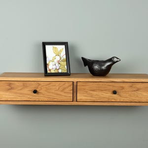 May include: A floating wooden shelf with two drawers, featuring a framed picture and a black bird-shaped decorative object. The shelf is made of light-colored wood and has black knobs on the drawers.