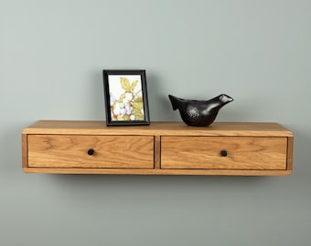 May include: A floating wooden shelf with two drawers, each featuring a small black circular knob. On top of the shelf sits a black rectangular picture frame containing a botanical illustration and a black ceramic bird figurine with textured wings.