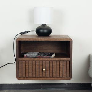 Può includere: Un comodino in legno montato a parete con finitura marrone scuro, dotato di un ripiano aperto e un cassetto singolo con listelli verticali e un piccolo pomello rotondo in ottone. Una lampada da tavolo nera con paralume bianco è posata sopra.