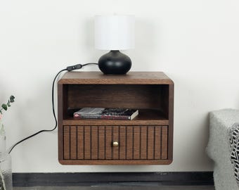 Può includere: Un comodino in legno montato a parete con finitura marrone scuro, dotato di un ripiano aperto e un cassetto singolo con listelli verticali e un piccolo pomello rotondo in ottone. Una lampada da tavolo nera con paralume bianco è posata sopra.