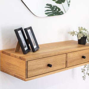 Op de afbeelding: Een licht houten zwevend plank met twee laden en zwarte knoppen. De plank is gemonteerd aan een witte muur en heeft twee zwarte fotolijsten en een plant in een pot erop.