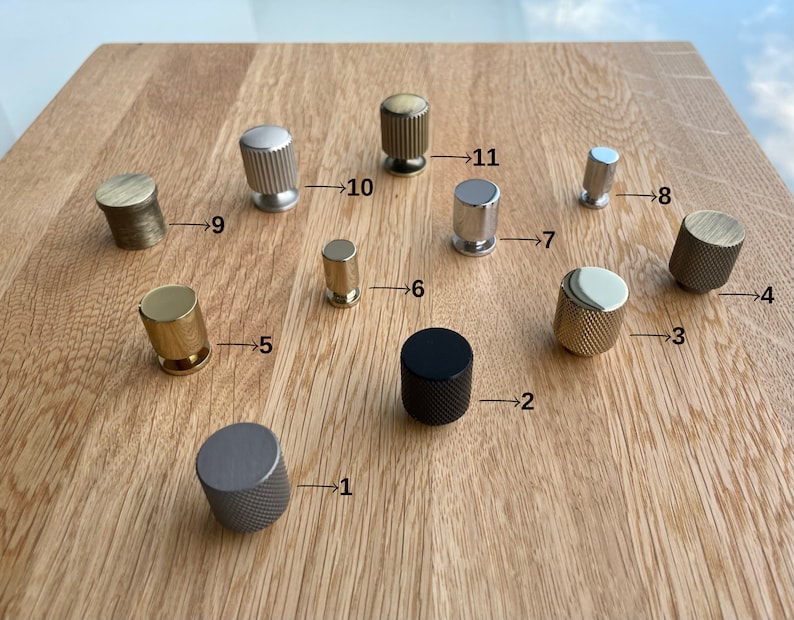 Puede incluir: Una colecci&oacute;n de once pomos de armario en varios acabados met&aacute;licos, incluyendo plata, oro y negro. Los pomos tienen diferentes texturas y formas, como dise&ntilde;os acanalados, moleteados y cil&iacute;ndricos. Est&aacute;n dispuestos sobre una superficie de madera clara.