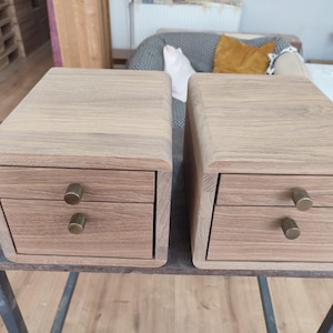 May include: Two wooden nightstands with two drawers each. The nightstands are made of light-colored wood with rounded edges and brass drawer pulls. The nightstands are on a dark surface.