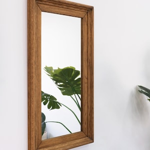 May include: A rectangular mirror with a brown wooden frame. The frame has a simple, slightly raised design. The mirror is reflecting a green plant.