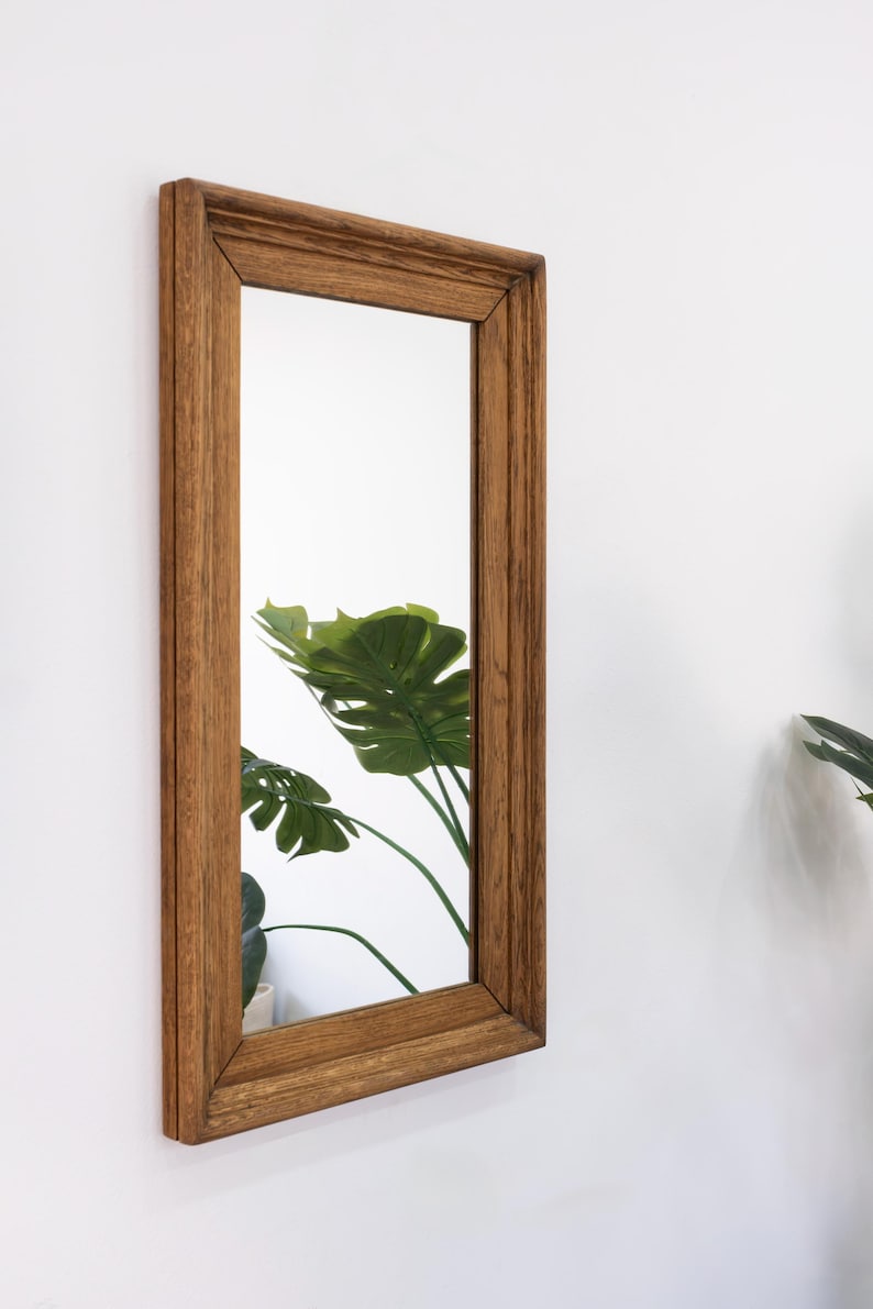 May include: A rectangular mirror with a brown wooden frame. The frame has a simple, slightly raised design. The mirror is reflecting a green plant.