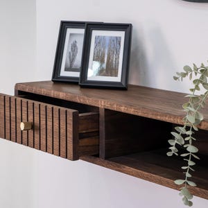 Può includere: Un ripiano a muro in legno marrone scuro con un cassetto e uno scaffale. Il cassetto è aperto e ha una manopola in ottone. Lo scaffale ha una piccola pianta in vaso. Ci sono due cornici per foto nere sullo scaffale.