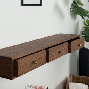 Puede incluir: Estante flotante de madera marr&oacute;n oscuro con tres cajones, cada uno con un pomo de lat&oacute;n. El estante est&aacute; montado en una pared blanca. Los cajones est&aacute;n parcialmente abiertos. Una soluci&oacute;n de almacenamiento moderna.