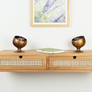 May include: A wall-mounted wooden floating shelf featuring two drawers with light-colored woven cane panels and circular cut-out handles. Two bronze-toned spherical candle holders sit on top, flanking a small, closed light green notebook.