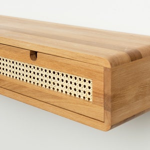 May include: A wall-mounted floating shelf made of light-toned wood with rounded edges. The front features two horizontal drawers, each inlaid with a rectangular panel of natural woven cane webbing and a small semi-circular cutout for opening.
