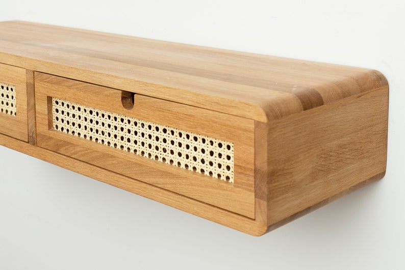 May include: A wall-mounted floating shelf made of light-toned wood with rounded edges. The front features two horizontal drawers, each inlaid with a rectangular panel of natural woven cane webbing and a small semi-circular cutout for opening.