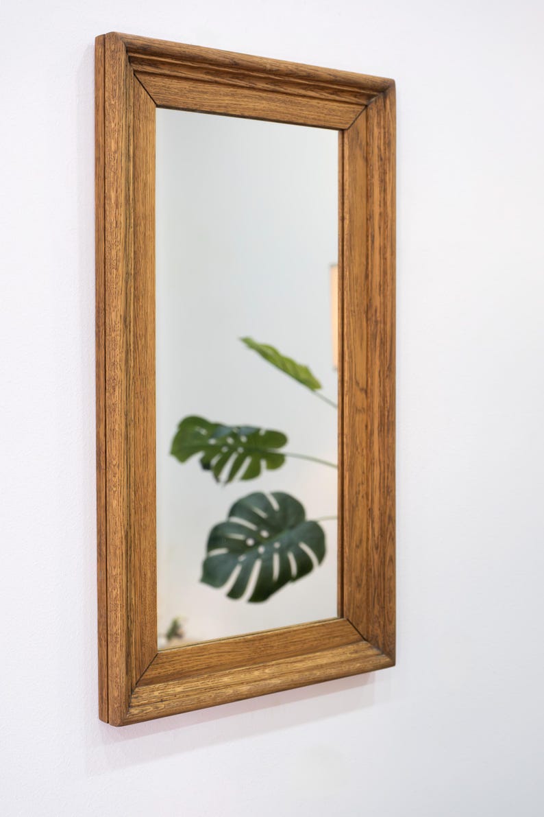 May include: A rectangular wooden framed mirror with a light brown finish. The mirror reflects a green leafy plant.