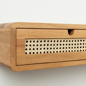 May include: A wall-mounted floating drawer unit crafted from light-toned wood with rounded corners. The front panel features a rectangular inset of woven cane webbing and a small semi-circular cutout at the top edge for opening the drawer.