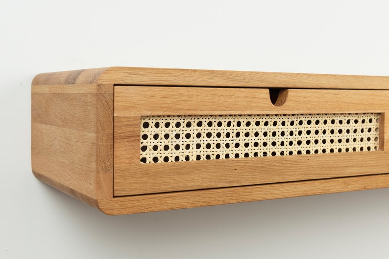 May include: A wall-mounted floating drawer unit crafted from light-toned wood with rounded corners. The front panel features a rectangular inset of woven cane webbing and a small semi-circular cutout at the top edge for opening the drawer.