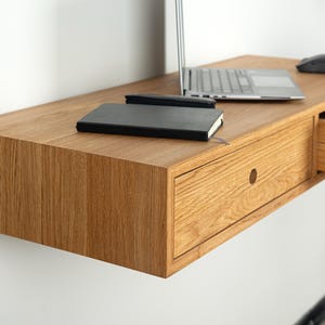 Peut inclure: Un bureau flottant en chêne clair fixé au mur avec une surface rectangulaire, un tiroir unique doté d'une poignée ronde en bois et un compartiment latéral entrouvert. Un ordinateur portable argenté, une souris noire et un carnet noir sont posés.