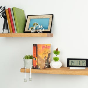 May include: Two light brown wooden floating shelves mounted on a white wall. The top shelf holds a black metal figurine, four books, and a framed picture. The bottom shelf holds a gray planter with dangling legs, two books, a white geometric pot, and a digital clock.