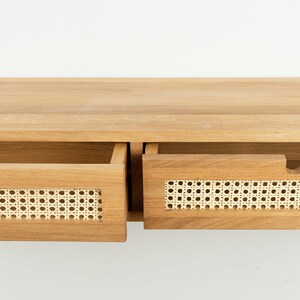 May include: A light brown wooden console table featuring two drawers with natural woven cane webbing inserts. Each drawer front includes a semi-circular cutout handle. The wood grain is visible across the flat top surface and drawer frames.
