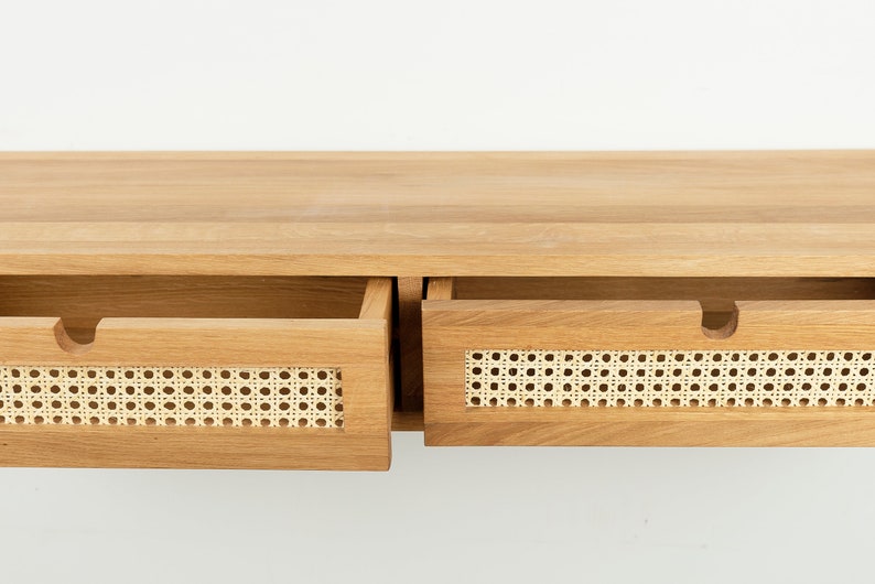 May include: A light brown wooden console table featuring two drawers with natural woven cane webbing inserts. Each drawer front includes a semi-circular cutout handle. The wood grain is visible across the flat top surface and drawer frames.