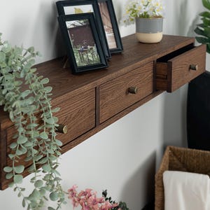 Puede incluir: Estante flotante de madera marr&oacute;n oscuro con dos cajones y tiradores de lat&oacute;n. Dos fotos enmarcadas y una peque&ntilde;a planta en maceta est&aacute;n encima. Tambi&eacute;n se ven una planta colgante y una cesta.
