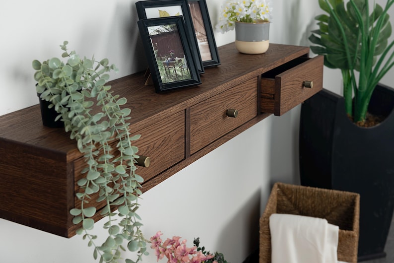 Puede incluir: Estante flotante de madera marr&oacute;n oscuro con dos cajones y tiradores de lat&oacute;n. Dos fotos enmarcadas y una peque&ntilde;a planta en maceta est&aacute;n encima. Tambi&eacute;n se ven una planta colgante y una cesta.