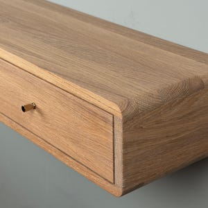 Könnte beinhalten: Ein wandmontiertes, schwebendes Regal mit einer natürlichen, hellbraunen Holzmaserung und abgerundeten Kanten. Das Möbelstück verfügt über eine Schubladenfront mit einem kleinen, zylindrischen, goldfarbenen Metallknopf.