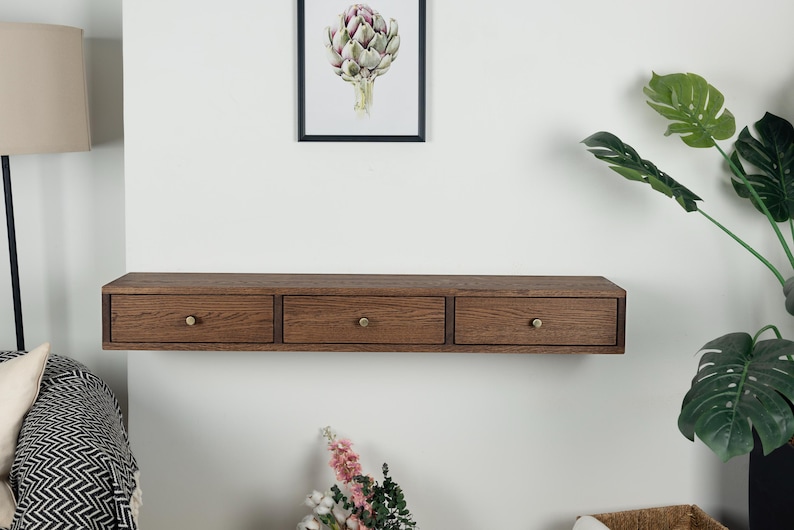 Puede incluir: Una estanter&iacute;a flotante de madera con tres cajones. La estanter&iacute;a es de color marr&oacute;n c&aacute;lido y tiene pomos de lat&oacute;n. Los cajones est&aacute;n espaciados uniformemente. La estanter&iacute;a est&aacute; montada en una pared blanca, con una impresi&oacute;n bot&aacute;nica enmarcada encima.