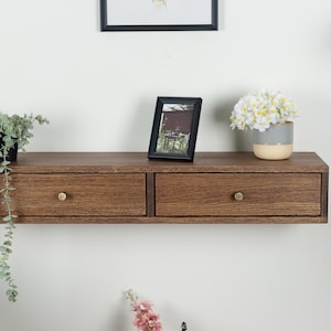 May include: A wooden floating shelf with two drawers and brass knobs. The shelf is mounted on a white wall and has a plant, a picture frame, and a vase of flowers on top.