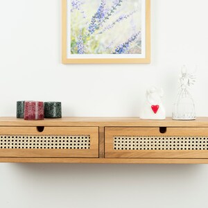 May include: A wall-mounted wooden floating shelf featuring two drawers with natural woven cane fronts. The shelf has a light brown wood finish and semi-circular cutouts for drawer handles. The top surface holds three small candles and two decorative figurines.