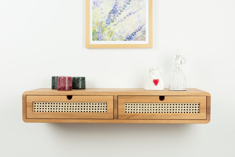 May include: A wall-mounted wooden floating shelf featuring two drawers with natural woven cane fronts. The shelf has a light brown wood finish and semi-circular cutouts for drawer handles. The top surface holds three small candles and two decorative figurines.