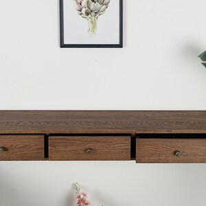 Puede incluir: Estante flotante de madera con tres cajones, cada uno con un pomo de lat&oacute;n. El estante es de color marr&oacute;n oscuro y est&aacute; montado en una pared blanca. Una impresi&oacute;n bot&aacute;nica enmarcada cuelga encima.