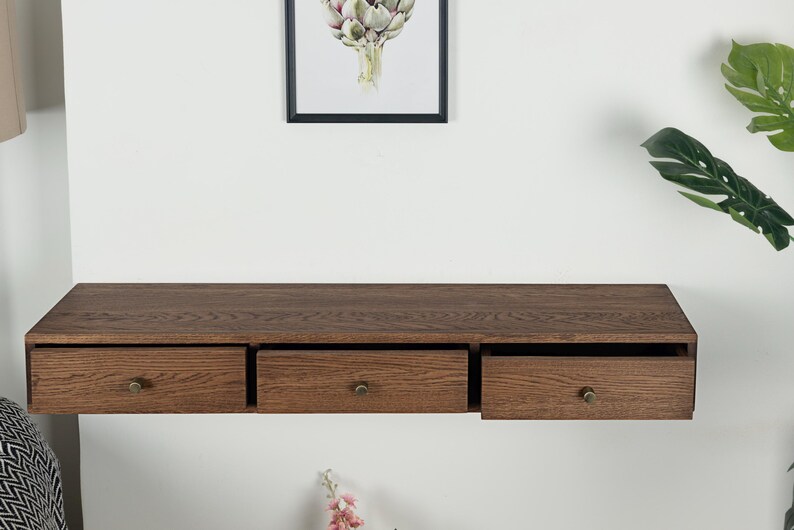 Puede incluir: Estante flotante de madera con tres cajones, cada uno con un pomo de lat&oacute;n. El estante es de color marr&oacute;n oscuro y est&aacute; montado en una pared blanca. Una impresi&oacute;n bot&aacute;nica enmarcada cuelga encima.