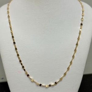 May include: A gold-coloured necklace featuring a repeating oval link pattern. The necklace is presented on a white mannequin bust, highlighting its length and design. The chain is slender and appears to be made of metal.