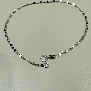 May include: A delicate silver anklet with a repeating pattern of small, flat, oval-shaped links. The anklet features a secure clasp closure. The jewelry is displayed on a light gray surface.