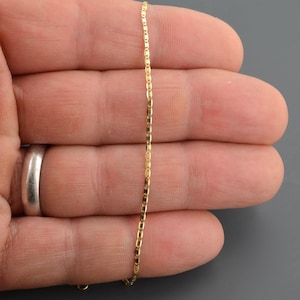 May include: A gold chain necklace with a box chain design. The chain is shown on a hand.