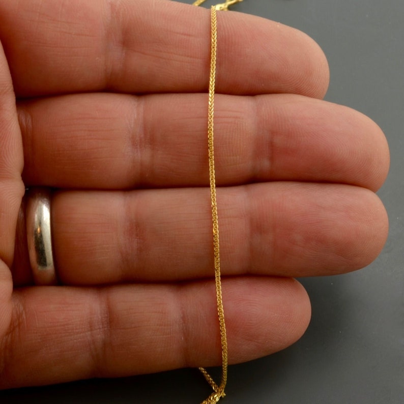 May include: A gold chain necklace with a delicate, braided design. The chain is shown on a person's hand.